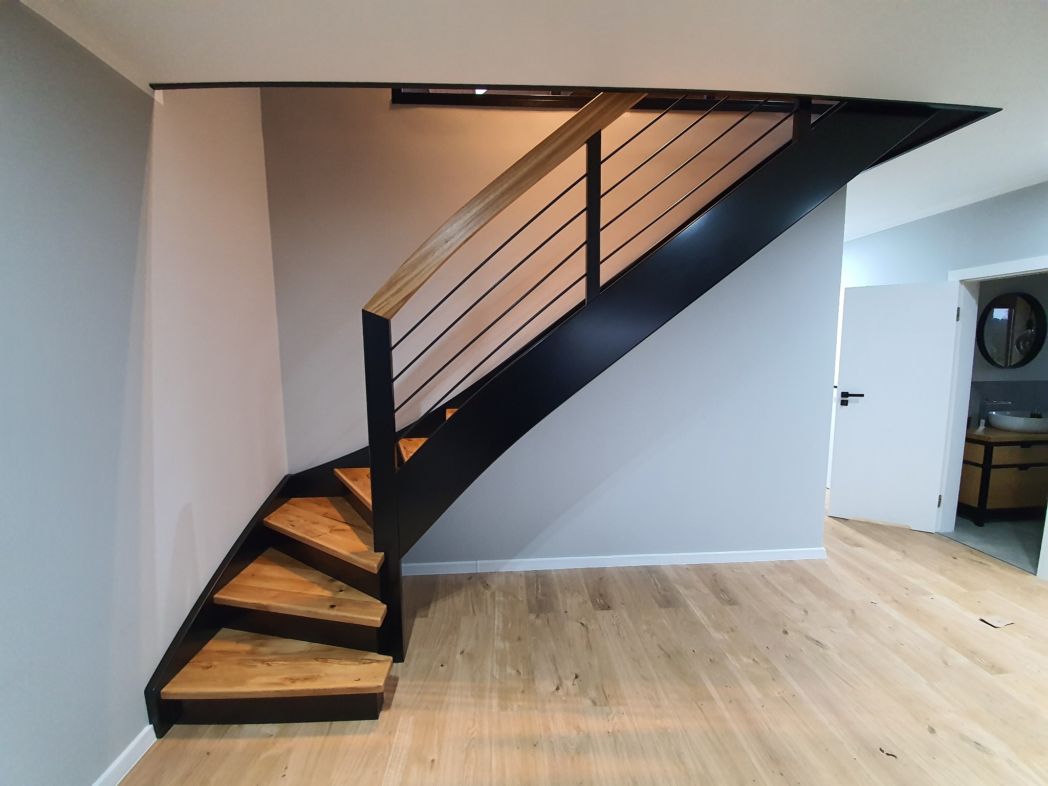 Nowoczesne schody z drewnianymi stopniami i czarną metalową konstrukcją, minimalistyczne balustrady z jasnego drewna, widok z dołu na jasną podłogę i drzwi do łazienki w tle.