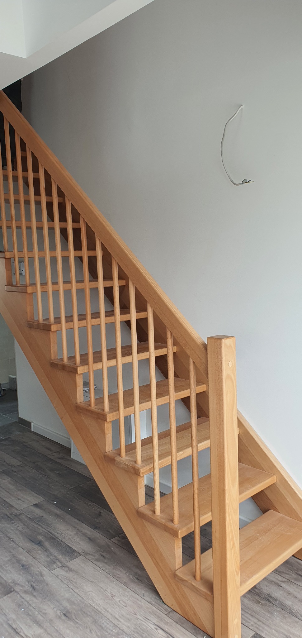 Jasne, drewniane schody z prostymi balustradami w jasnym wnętrzu, widoczny wystający kabel ze ściany.