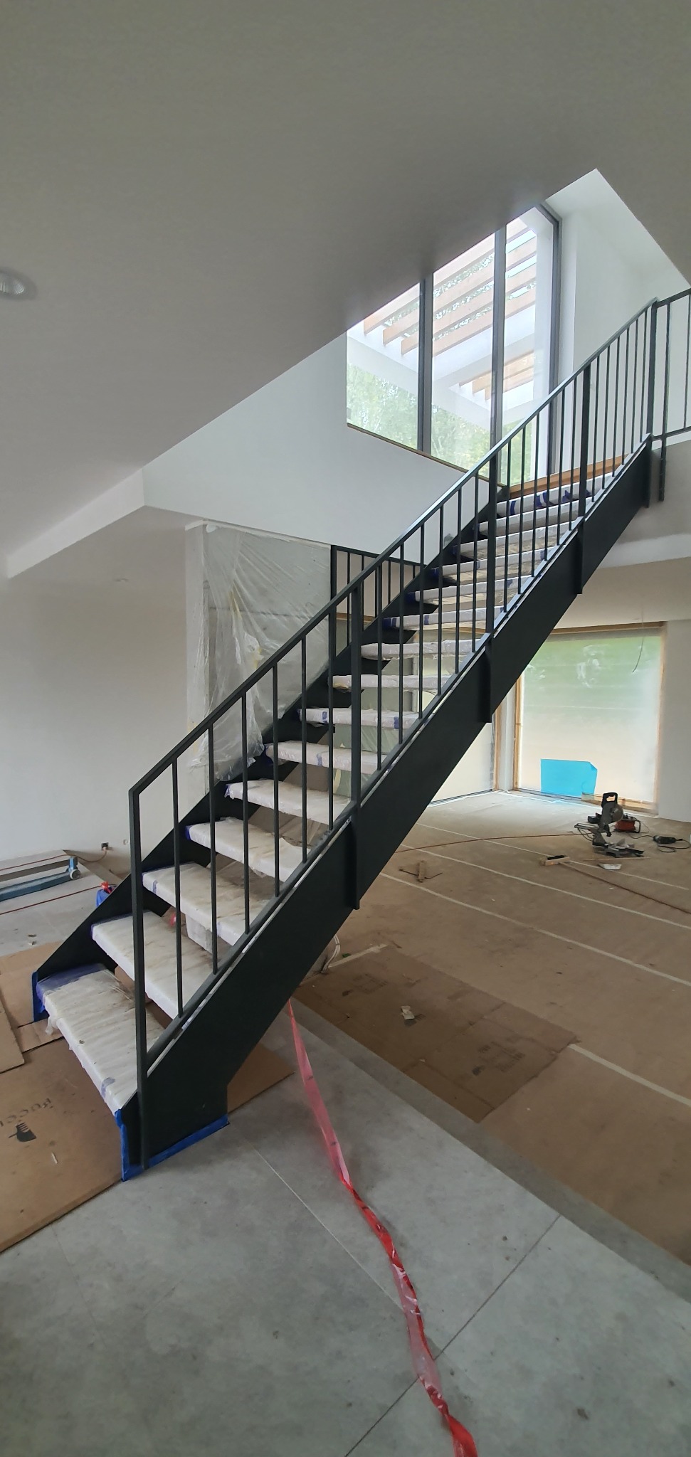 Nowoczesne schody z czarną metalową balustradą i schodami zabezpieczonymi folią, widoczne podczas montażu w jasnym wnętrzu z dużym oknem.
