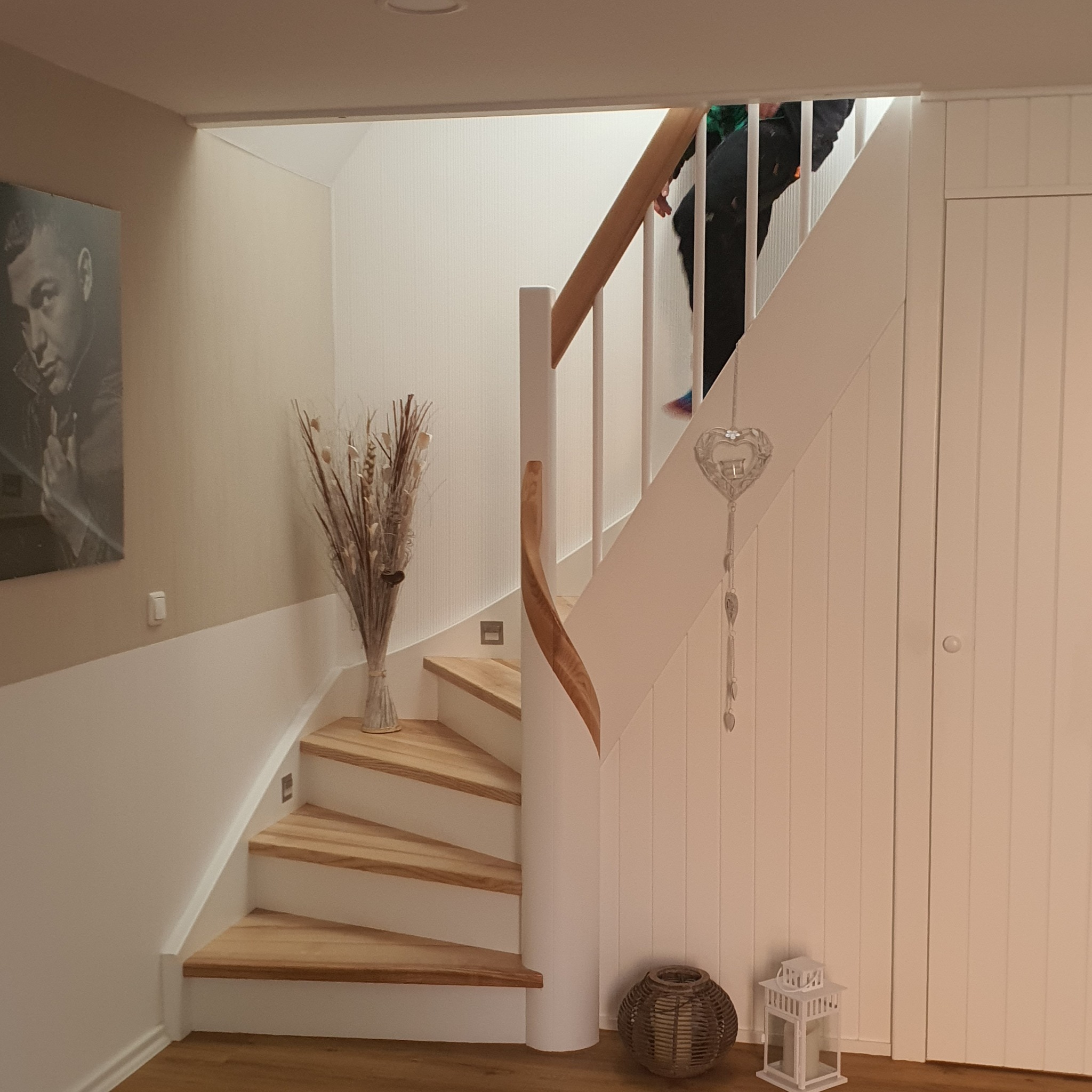 Białe schody z drewnianymi stopniami i poręczą, ozdobione wazonem z suszonymi kwiatami i dekoracją w kształcie serca, widoczne fragmenty osoby wchodzącej po schodach.