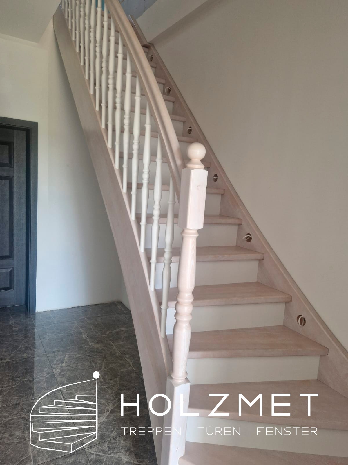 Drewniane schody o jasnej kolorystyce z białymi balustradami, widok z dołu, na podłodze błyszczące płytki imitujące marmur, w tle ciemne drzwi.