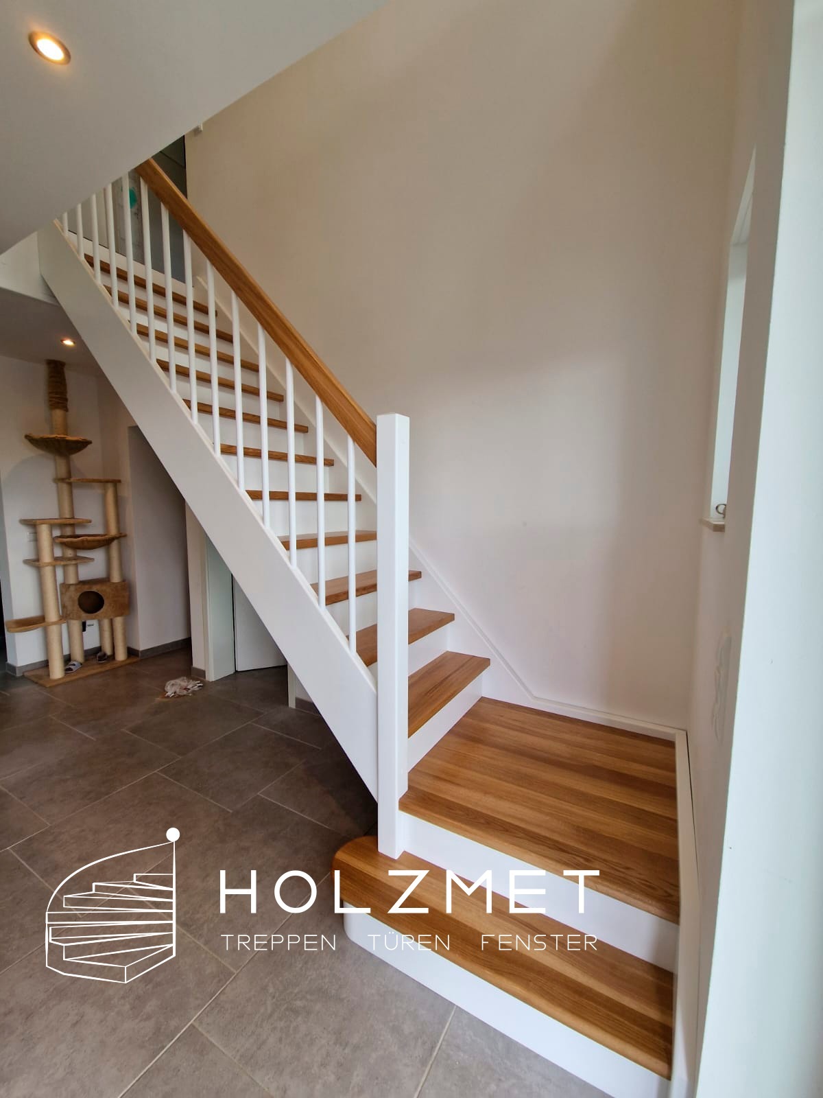 Nowoczesne schody z drewnianymi stopniami i białą balustradą w jasnym wnętrzu, widoczny drapak dla kota w tle.