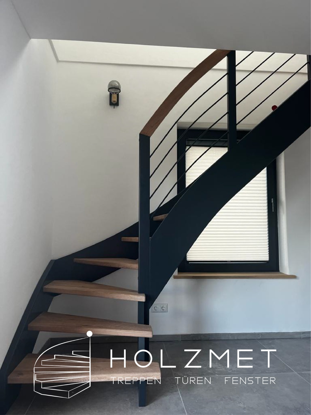 Spiralne schody z drewnianymi stopniami i czarną metalową konstrukcją, widoczne od dołu, z oknem w tle i lampą na ścianie. Na pierwszym planie logo firmy.