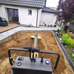 San Ran - Operator z perspektywy pierwszej osoby obsługuje małą, żółtą maszynę do zagęszczania gruntu na przygotowanym terenie pod kostkę brukową. W tle widoczny dom z szarą dachówką i zadbany ogród.