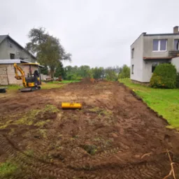 Żółta mini koparka na działce z przygotowanym terenem pod budowę, widoczne ślady opon w błocie, w tle domy mieszkalne i drzewa, pochmurne niebo.