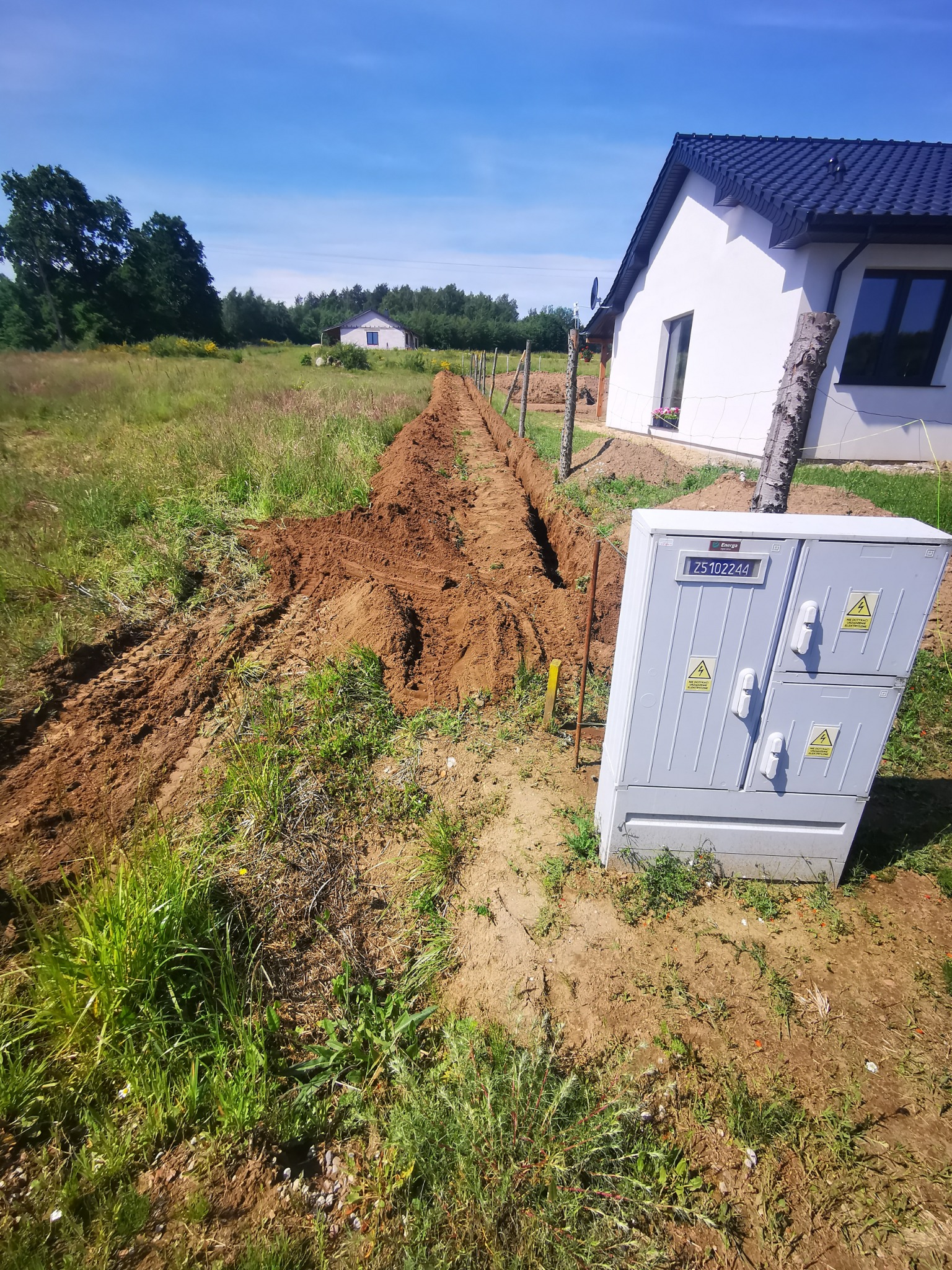 Wykopany rów w ziemi wzdłuż ogrodzenia, z widoczną skrzynką elektryczną i nowym domem w tle, wykonany w słoneczny dzień.