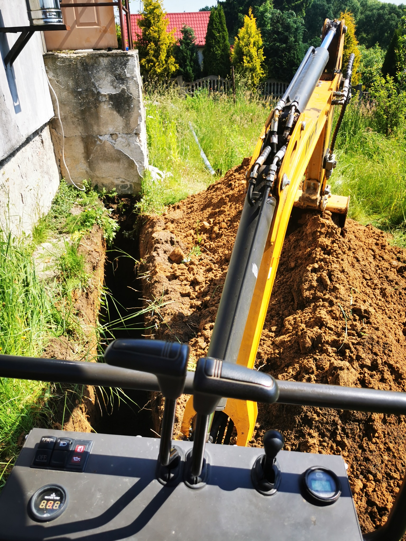 Żółta koparka z widokiem z kabiny, wykonująca wykop wąskoprzestrzenny w ziemi obok fundamentów budynku w słoneczny dzień.