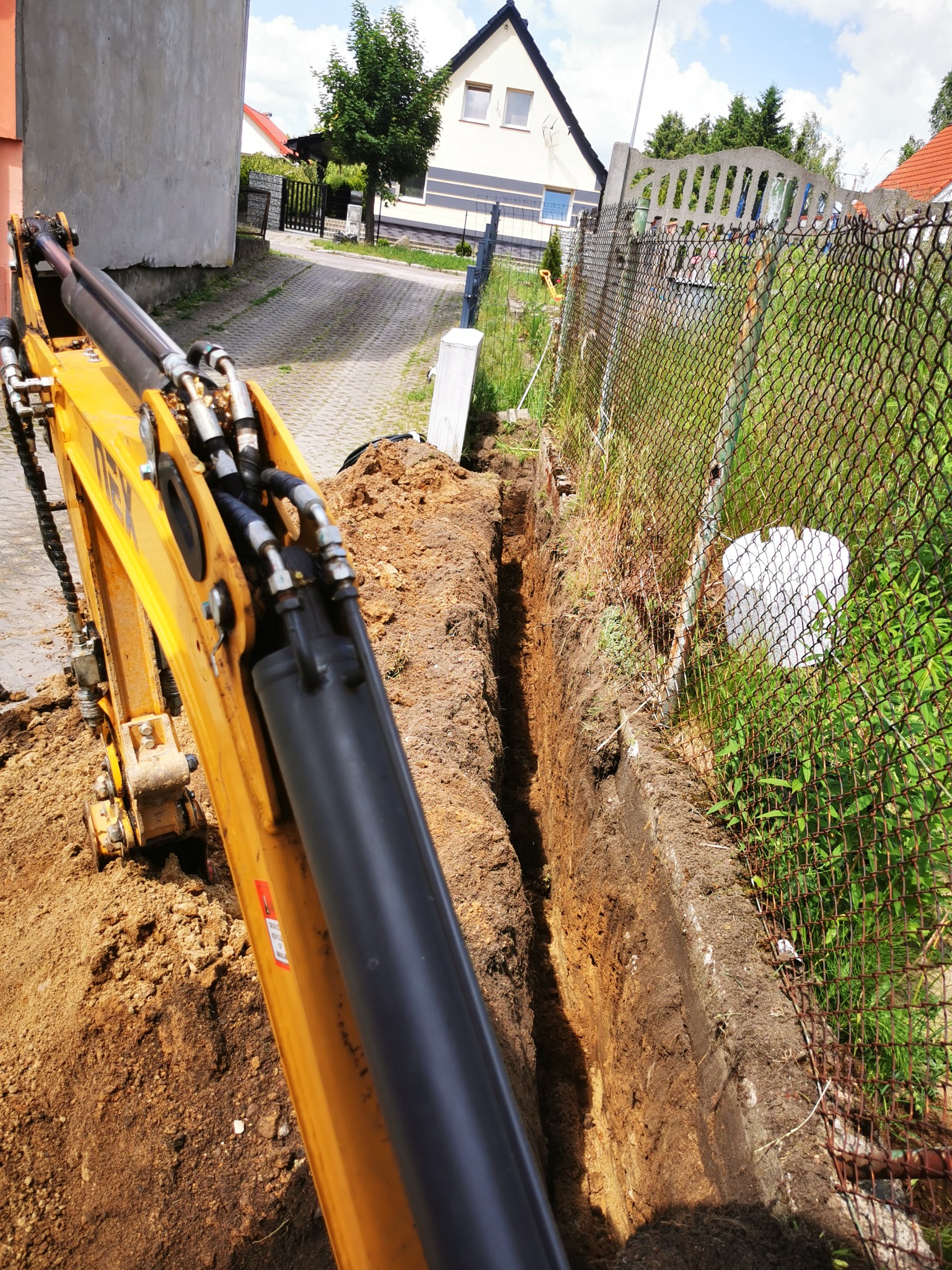 Żółta koparka wykopuje wąski rów w ziemi wzdłuż ogrodzenia z siatki, w tle widoczny dom z dwuspadowym dachem i podjazd.