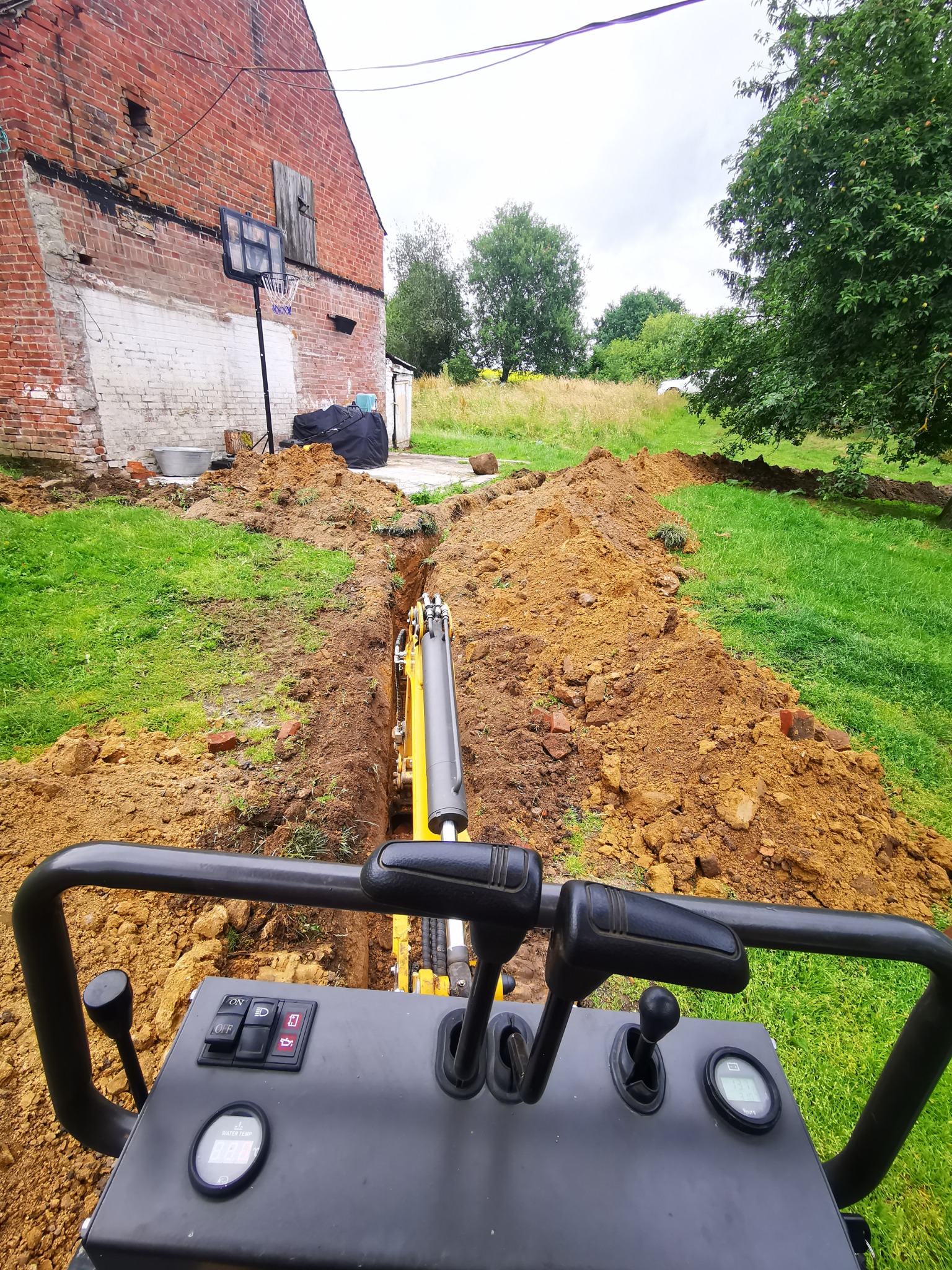 Wąski wykop pod instalacje wykonany minikoparką na trawiastym terenie z widocznym starym budynkiem z czerwonej cegły i koszem do koszykówki w tle.