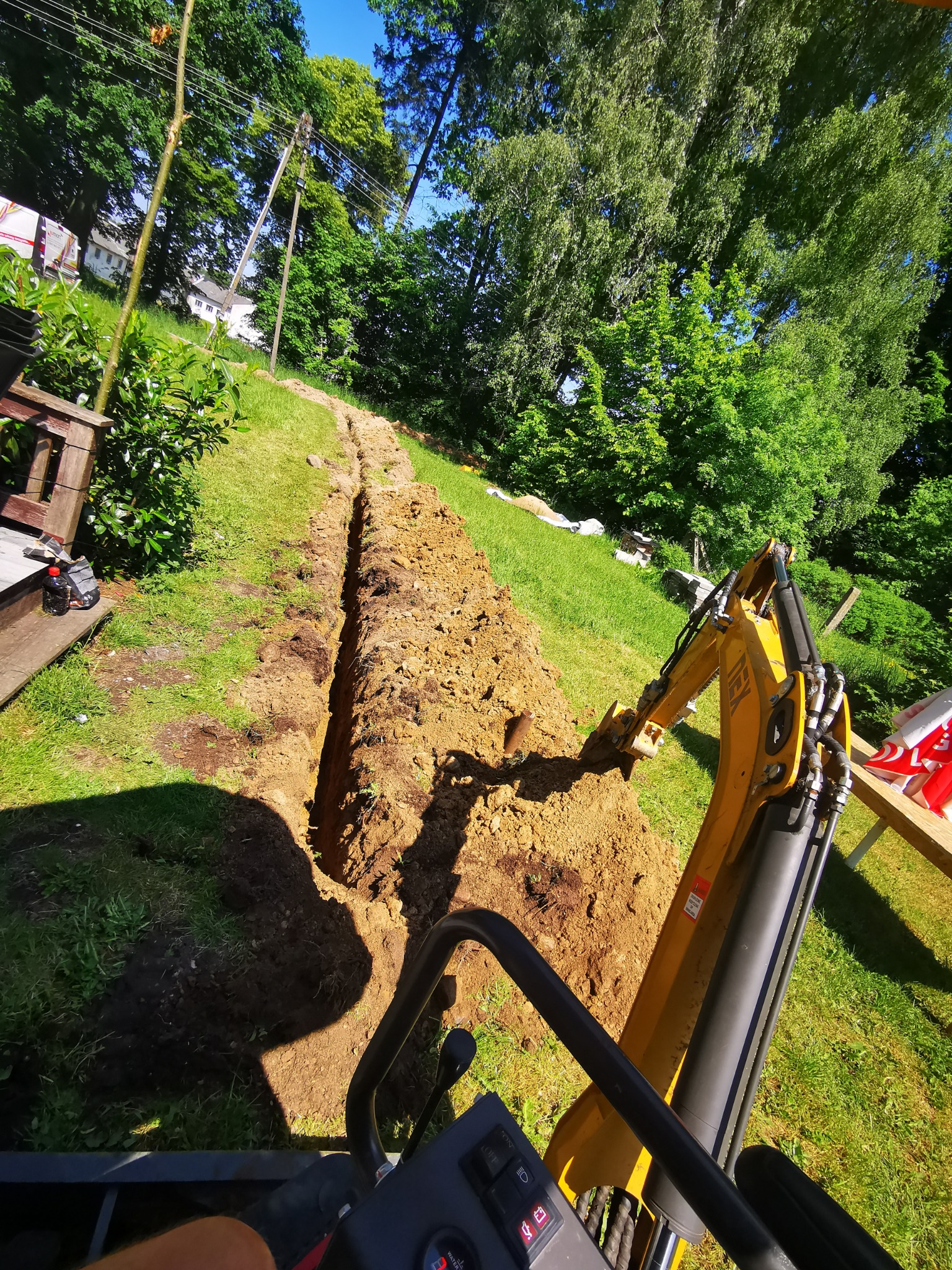 Wykopany wąski rów w trawie w Połczynie-Zdroju, wykonany żółtą mini koparką, widok z perspektywy operatora, zielona trawa i drzewa w tle.