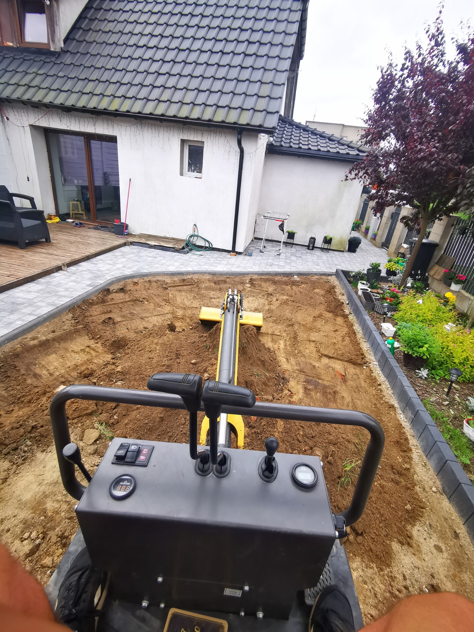 Operator z perspektywy pierwszej osoby obsługuje małą, żółtą maszynę do zagęszczania gruntu na przygotowanym terenie pod kostkę brukową. W tle widoczny dom z szarą dachówką i zadbany ogród.