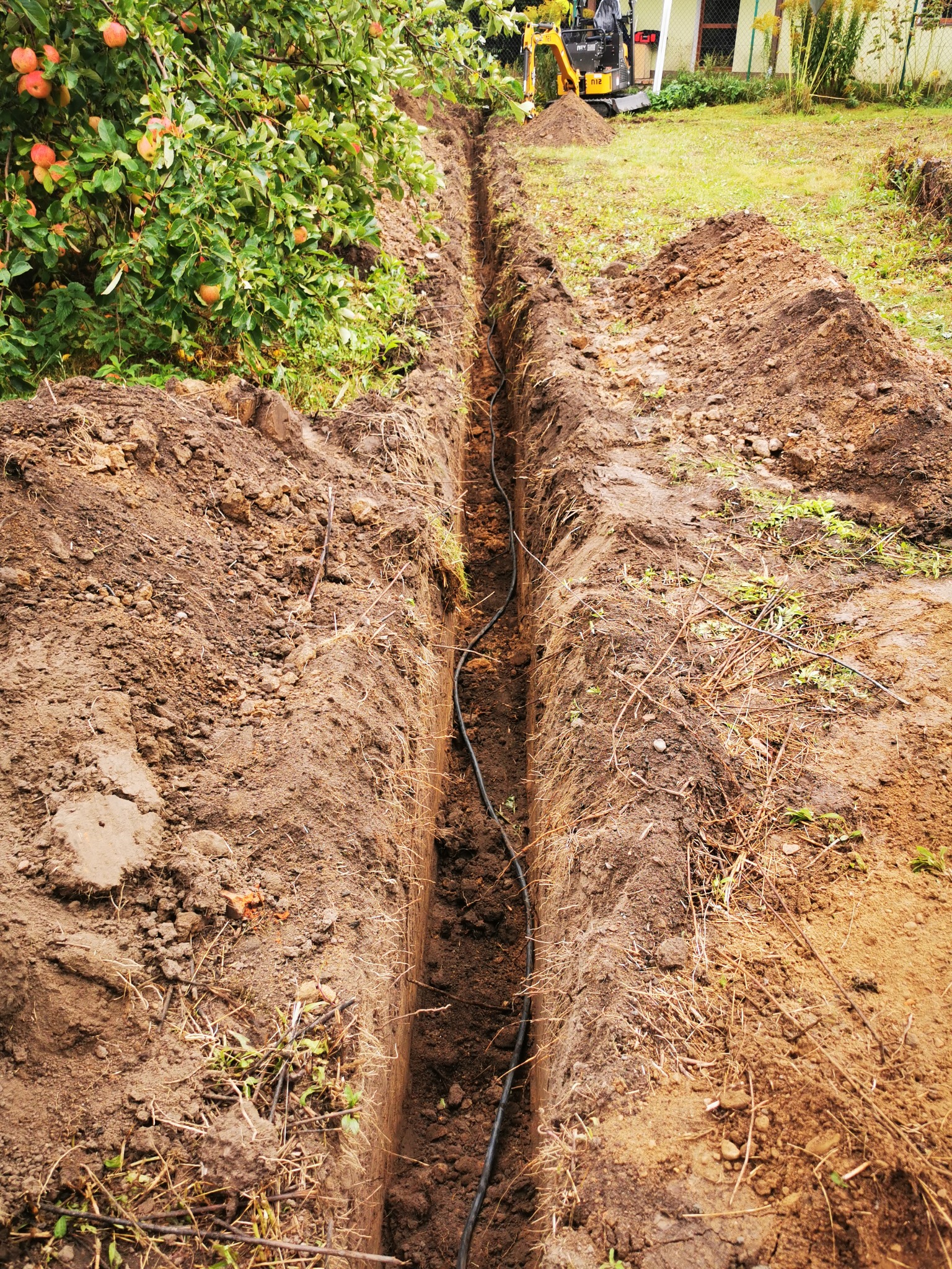 Wykopany wąski rów w ziemi z ułożonym kablem, widoczny mały żółty koparka w tle, obok rosnące jabłonie