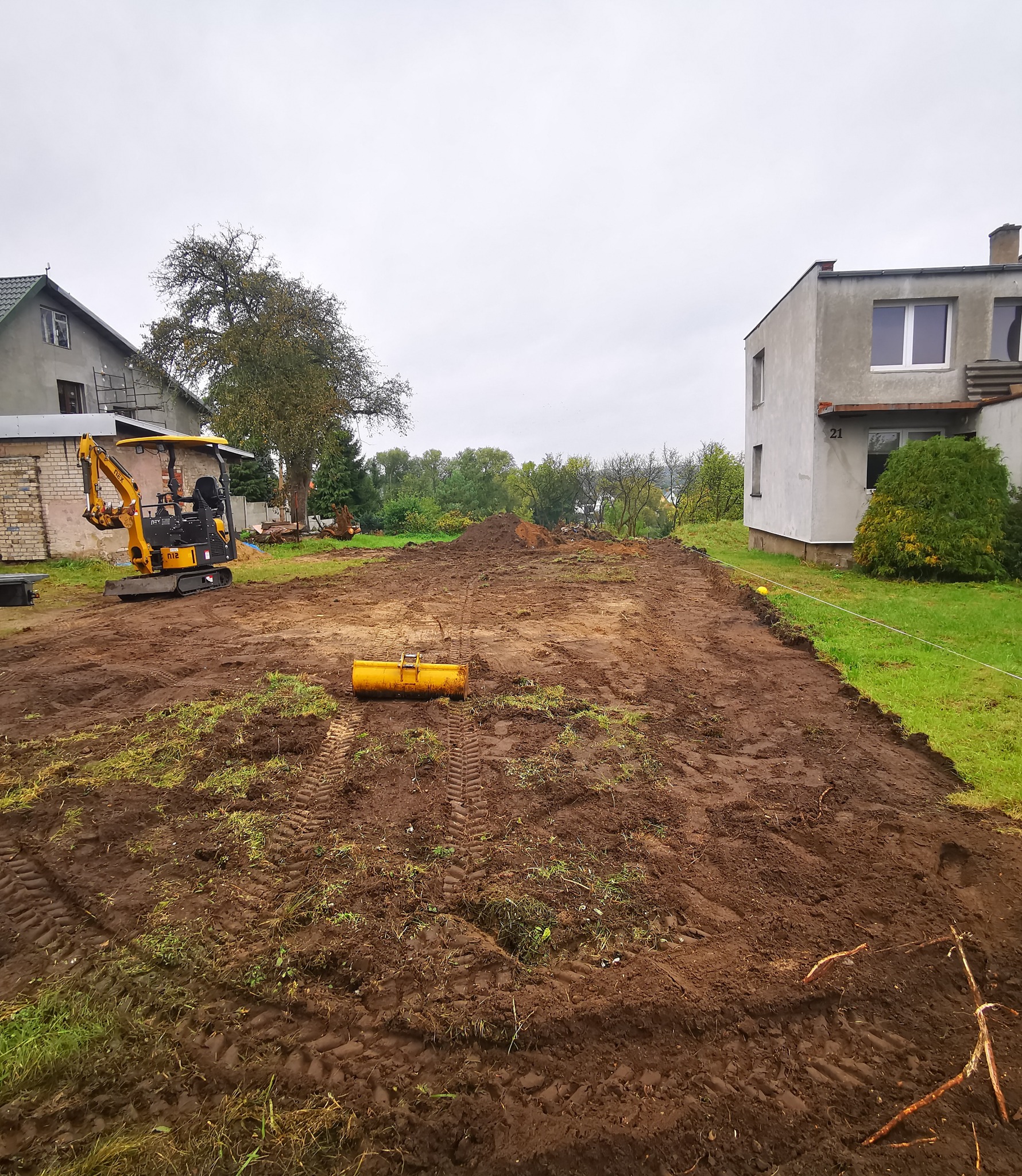 Żółta mini koparka na działce z przygotowanym terenem pod budowę, widoczne ślady opon w błocie, w tle domy mieszkalne i drzewa, pochmurne niebo.