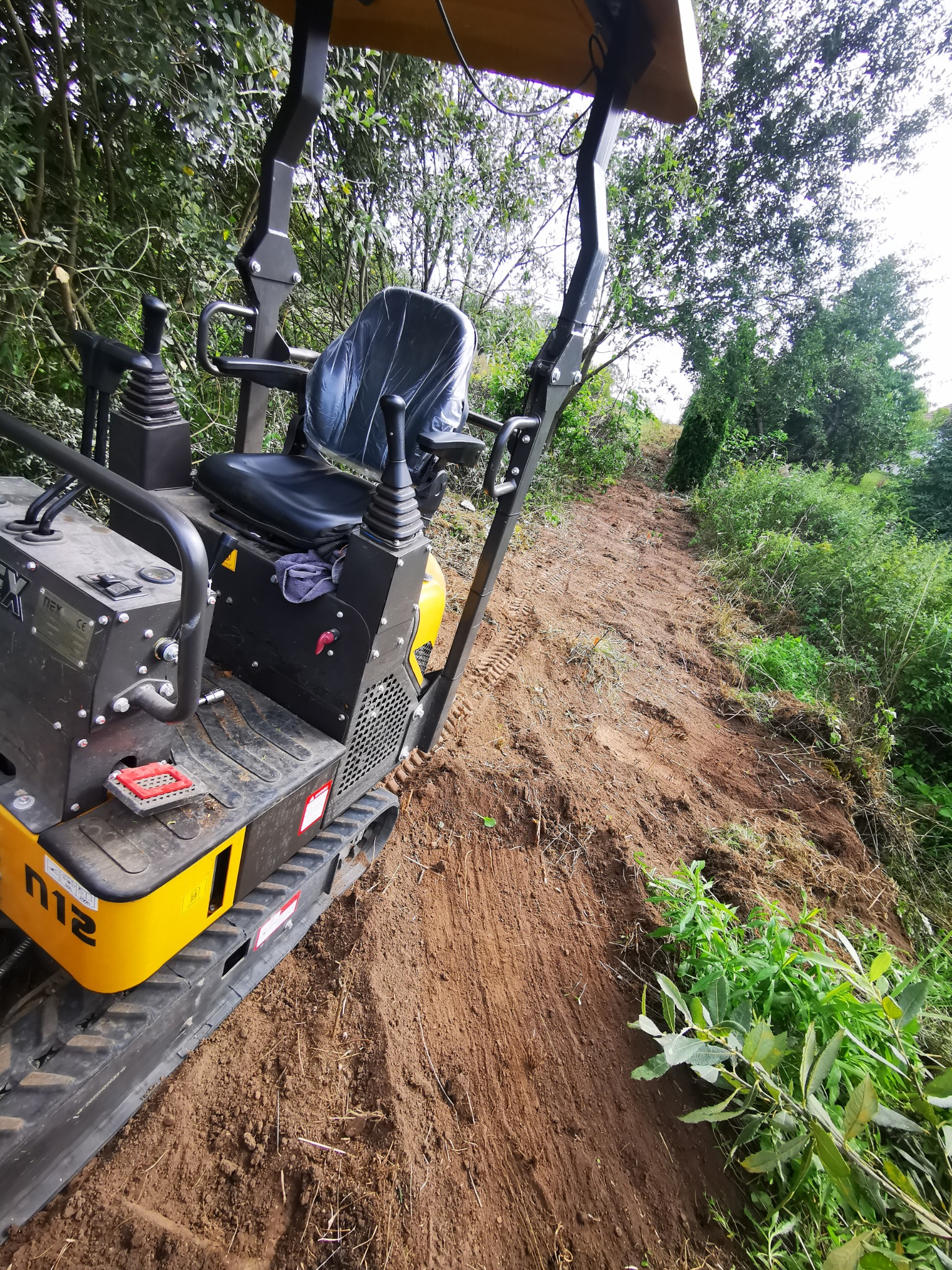 Żółto-czarna mini koparka na gąsienicach, widoczna z boku, stoi na wyrównanym, ziemistym terenie z pozostałościami roślinności, w tle zielone drzewa i krzewy.