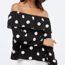 Czarno-biała bluzka w grochy z odkrytymi ramionami i falbanami, noszona przez modelkę z delikatnym makijażem.