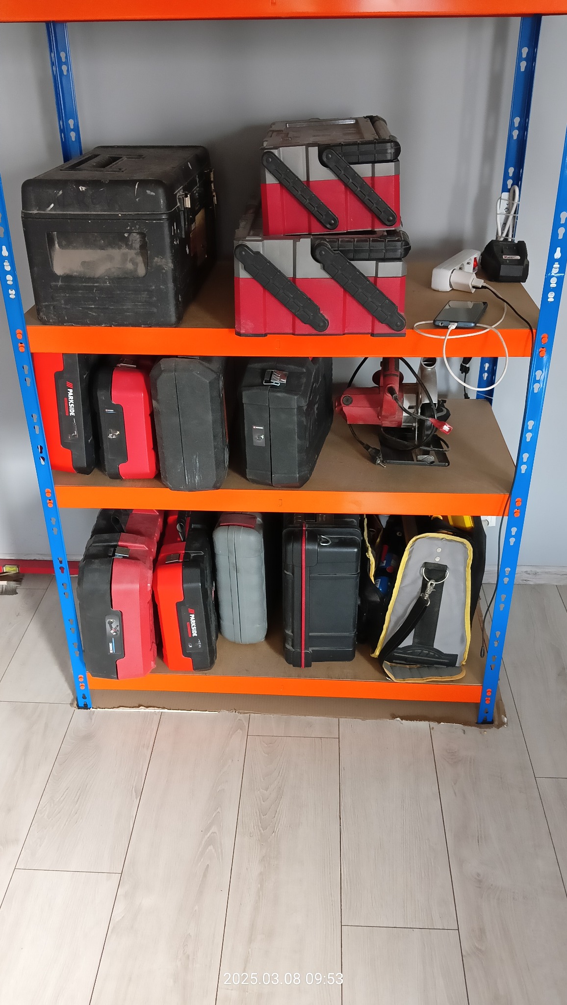 Regał warsztatowy z narzędziami i akcesoriami w skrzynkach, widoczna piła tarczowa, w tle jasne panele podłogowe.