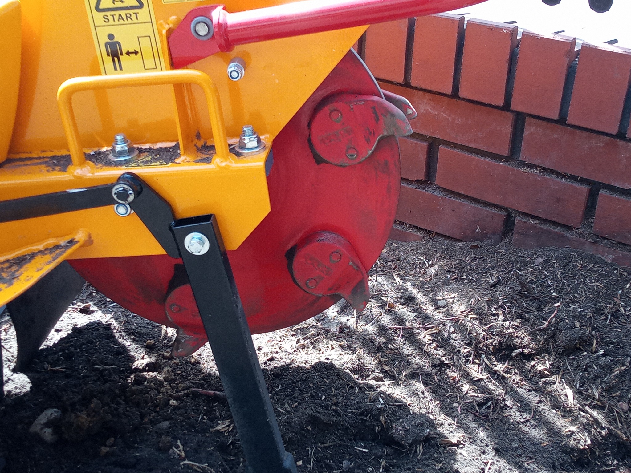 Fragment glebogryzarki z czerwonymi ostrzami, żółtą obudową i czarnym wspornikiem, pracującej na przygotowanej ziemi obok muru z czerwonej cegły.