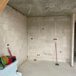 Surowy stan wnętrza z instalacją elektryczną w peszlach na ścianach z betonu komórkowego, widoczne puszki pod gniazdka i włączniki, przewody zwisające z surowego betonowego stropu, oraz narzędzia...