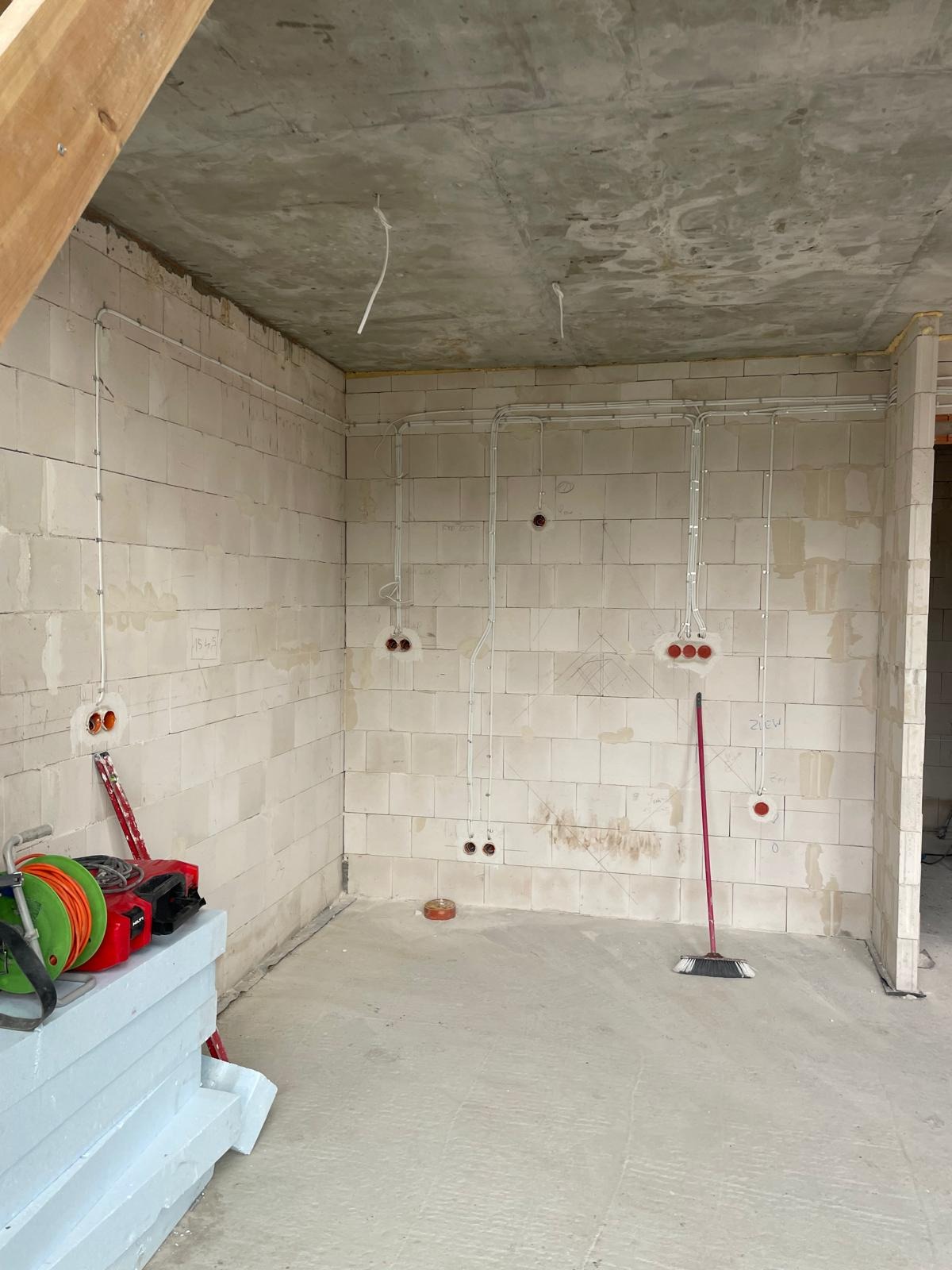 Surowy stan wnętrza z instalacją elektryczną w peszlach na ścianach z betonu komórkowego, widoczne puszki pod gniazdka i włączniki, przewody zwisające z surowego betonowego stropu, oraz narzędzia...