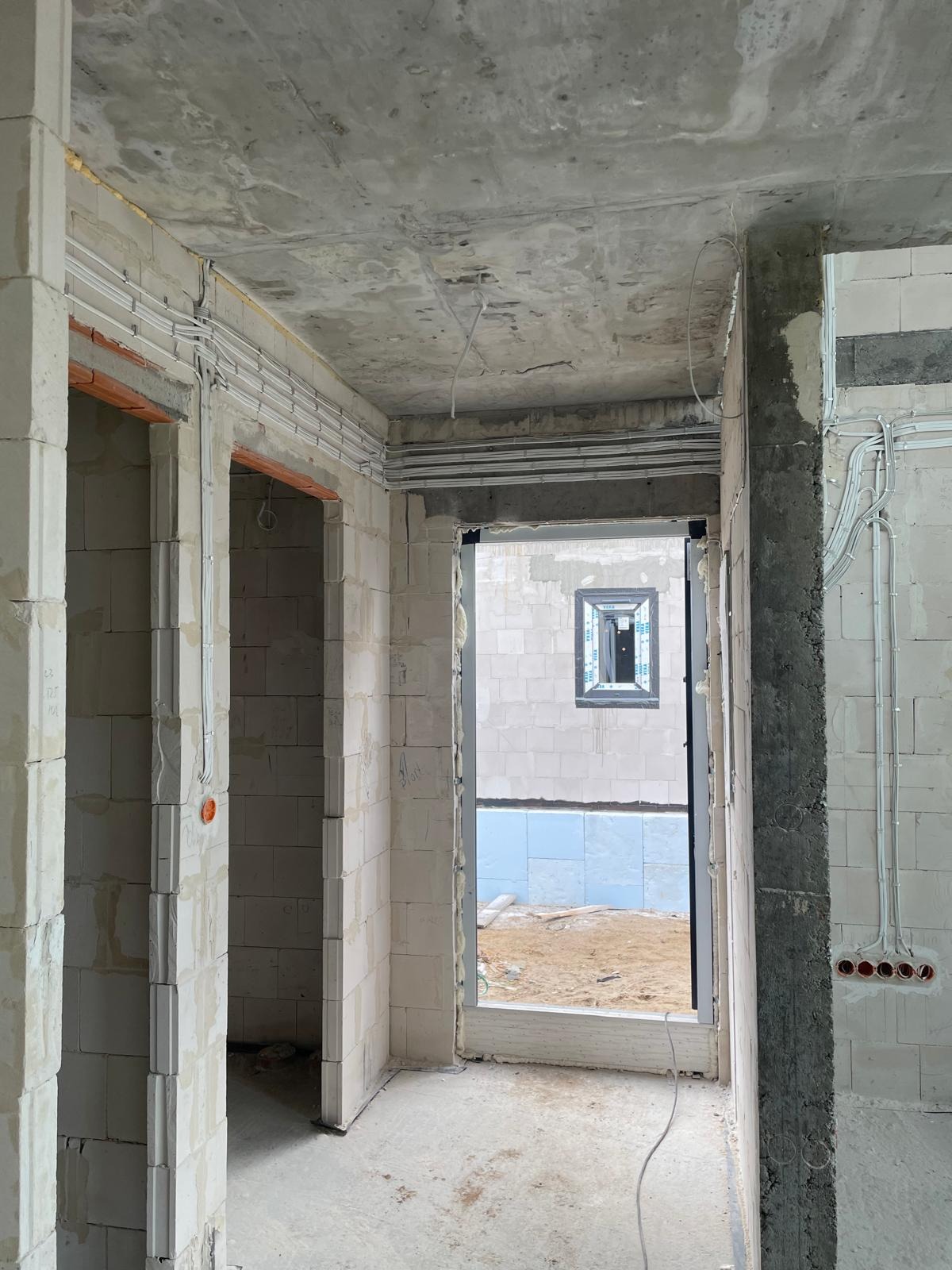 Surowy stan wnętrza budynku w trakcie prac instalacyjnych: widoczne białe ściany z betonu komórkowego, ułożone rury instalacji elektrycznej w peszlach, otwory drzwiowe i okienne, szary betonowy sufit.