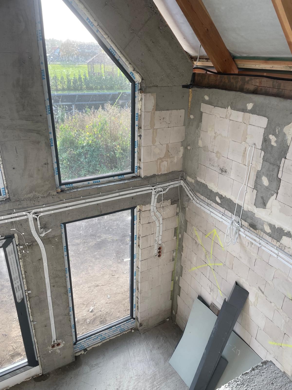 Surowy stan wnętrza poddasza z widocznymi oknami, instalacją elektryczną w peszlach i fragmentami nieotynkowanych ścian z betonu komórkowego z naniesionymi oznaczeniami.