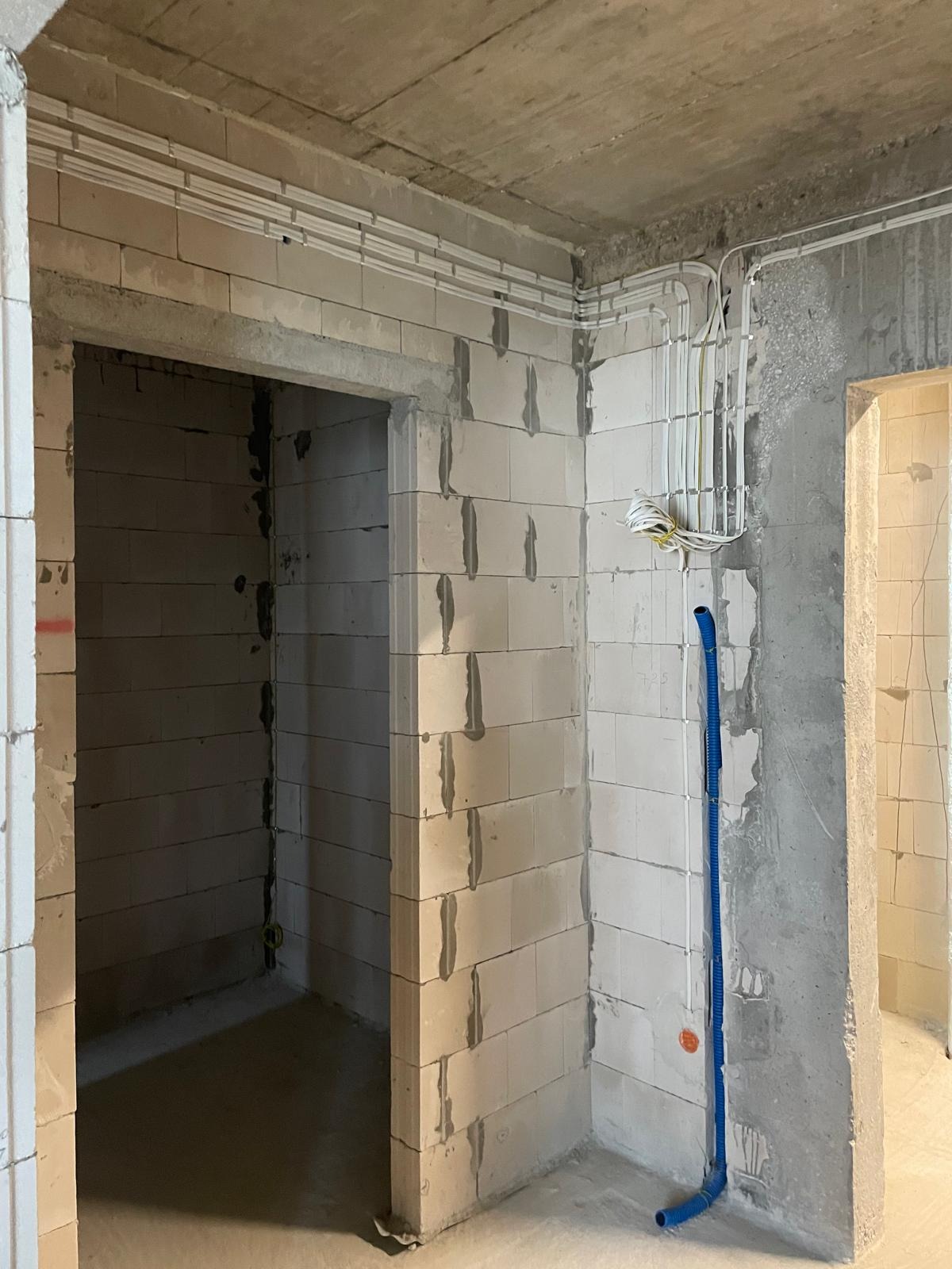 Surowy stan wnętrza z widocznymi przewodami elektrycznymi ułożonymi w korytkach na ścianach z betonu komórkowego, nieotynkowane ściany i sufit, pionowy niebieski przewód instalacyjny przy ścianie.