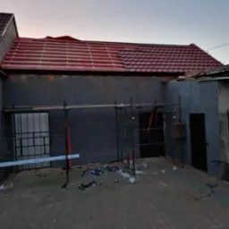 Częściowo pokryty dach z czerwonej blachodachówki, widoczne rusztowania i śmieci na podwórku, budynek w trakcie prac remontowych.