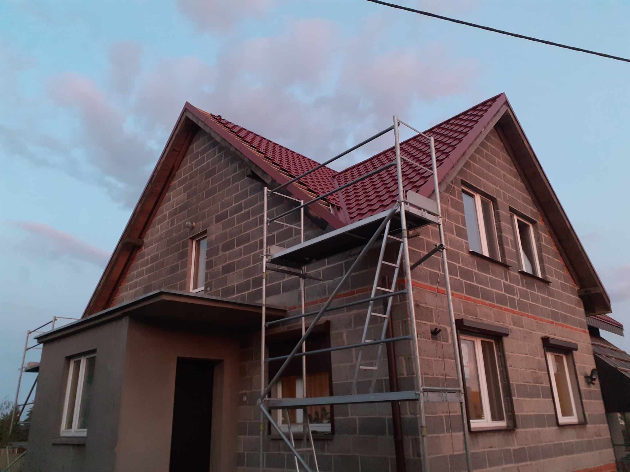 Dom w trakcie budowy z częściowo ułożonym czerwonym dachem, widoczne rusztowanie i bloczki betonowe.