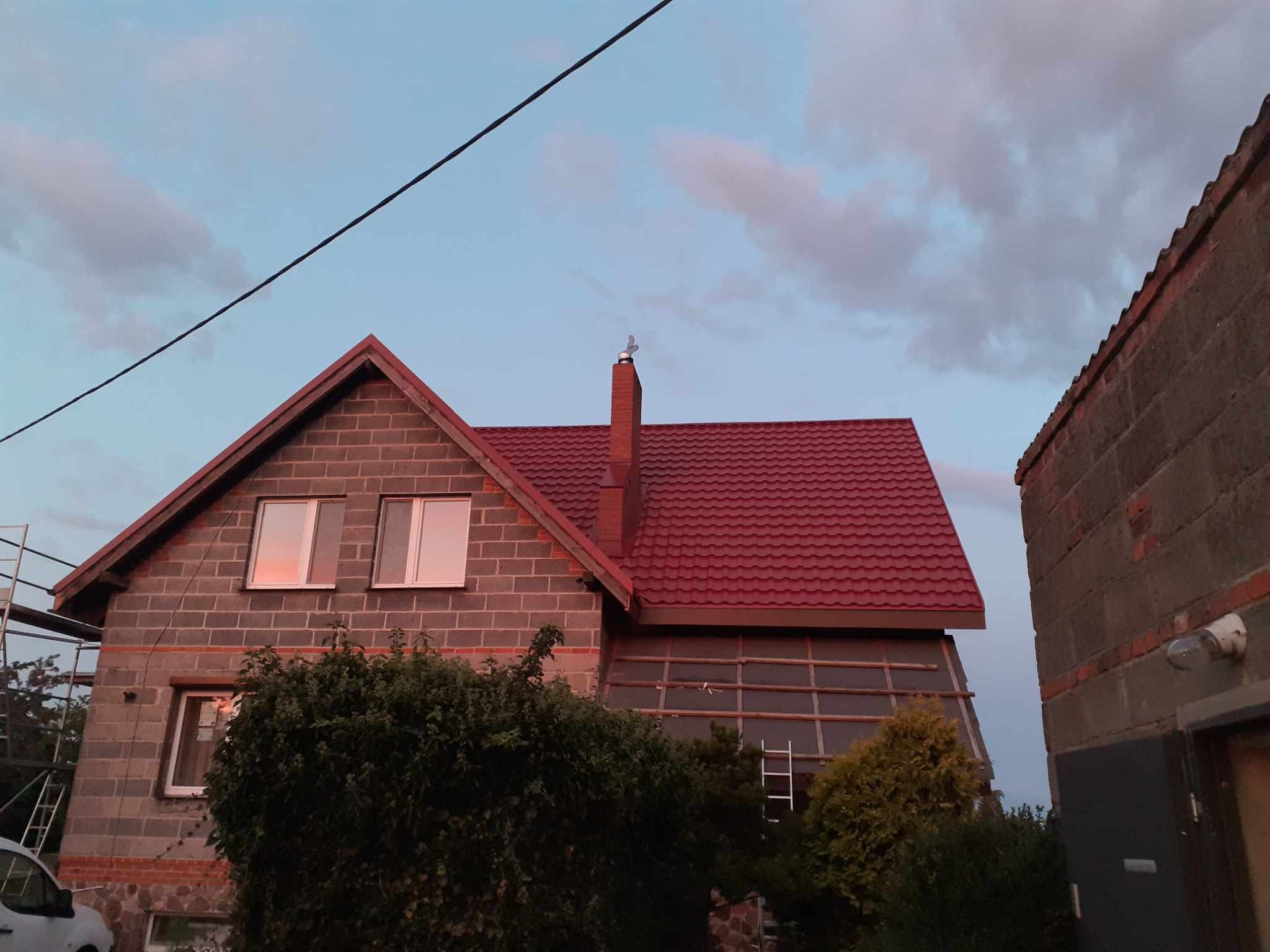 Dom w trakcie prac dekarskich: czerwona blachodachówka na nowym dachu, widoczne rusztowania i przygotowane podłoże pod ocieplenie poddasza, komin z ozdobną wiatrowskazą.