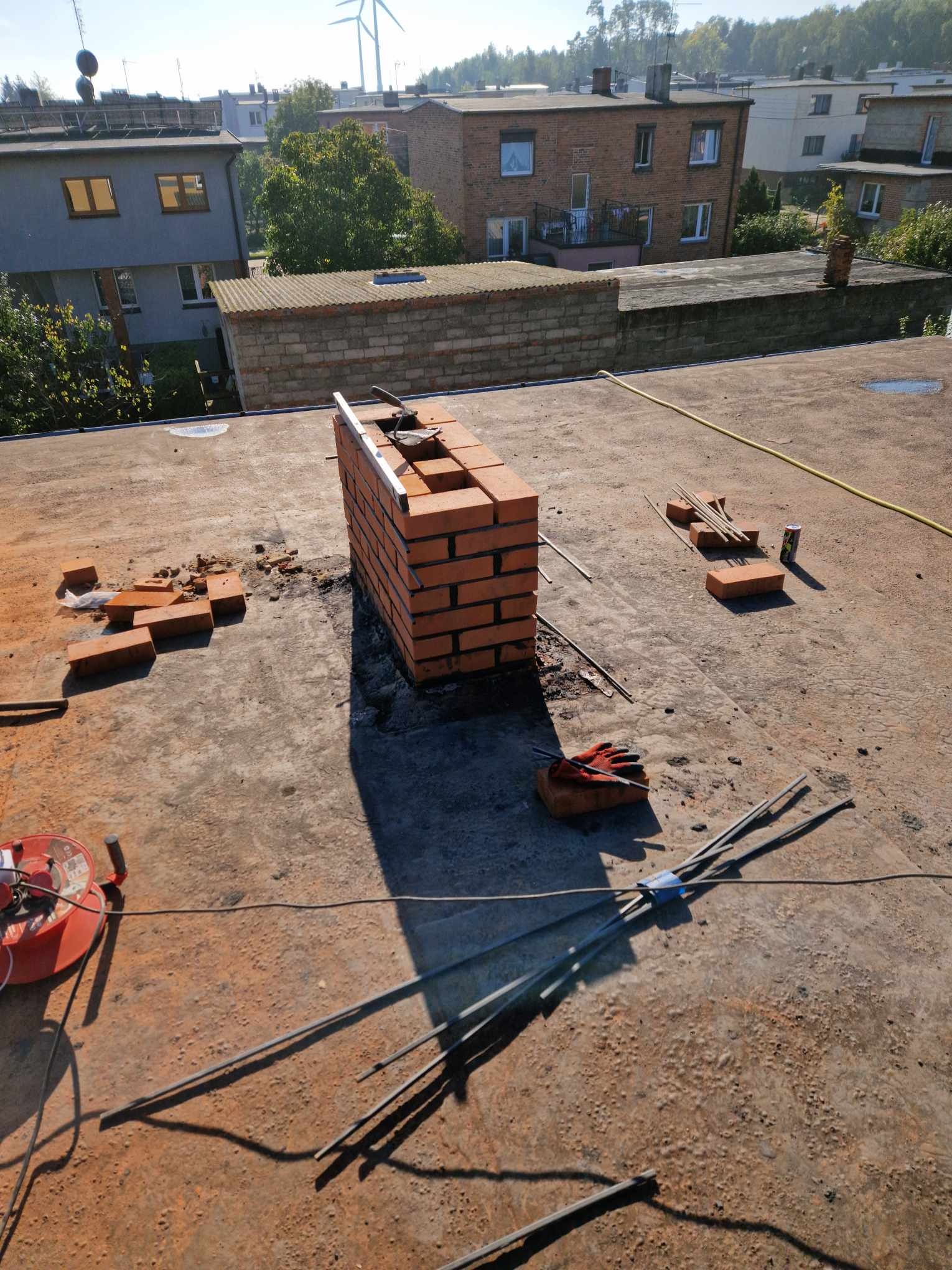 Wznoszenie komina z czerwonej cegły na płaskim dachu, widoczne narzędzia, fragmenty cegieł i rękawice robocze, w tle zabudowa miejska i turbiny wiatrowe.