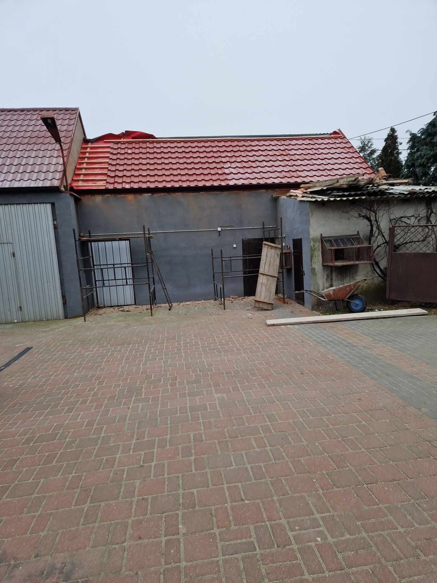 Częściowo wymieniony dach z czerwonej dachówki na budynku gospodarczym, widoczne rusztowania, gruz i przygotowane deski, na pierwszym planie bruk z czerwonej kostki.