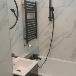 Nowoczesna łazienka z marmurowymi płytkami, czarną armaturą, grzejnikiem drabinkowym i okrągłym lustrem z podświetleniem LED.