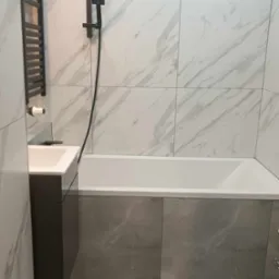 Nowoczesna łazienka z wanną obudowaną szarymi płytkami, kabiną prysznicową w kolorze czarnym, umywalką z szarą szafką i grzejnikiem drabinkowym.