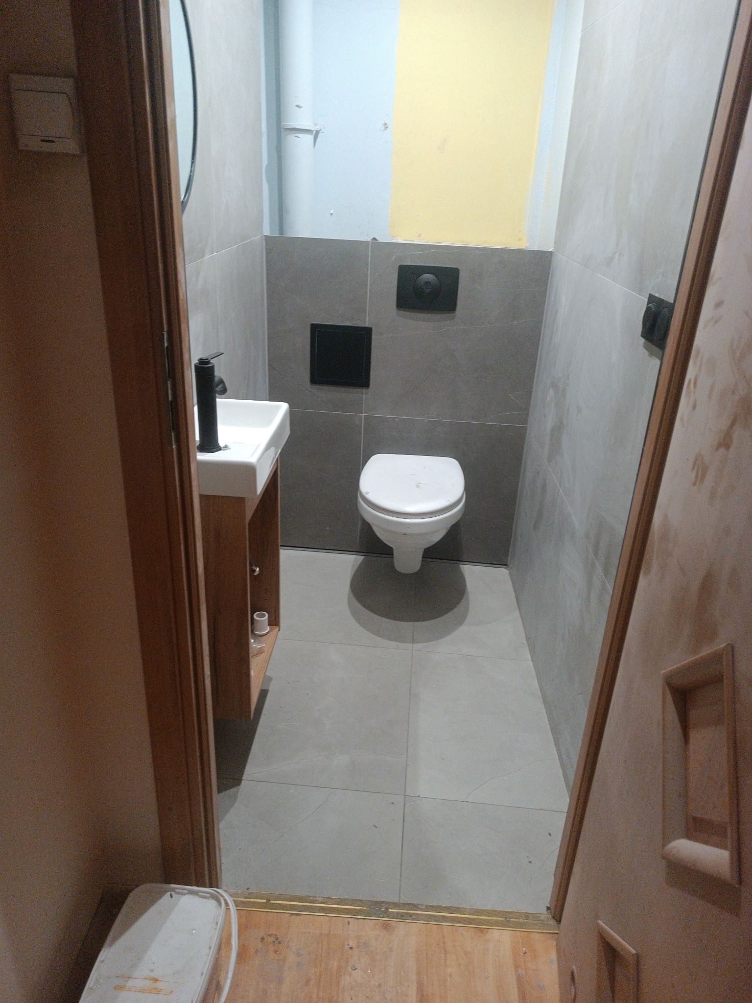 Mała łazienka w trakcie remontu: szare płytki na ścianach i podłodze, wisząca toaleta, umywalka z szafką z jasnego drewna i czarną baterią, widoczny biały pojemnik i niedokończone malowanie...