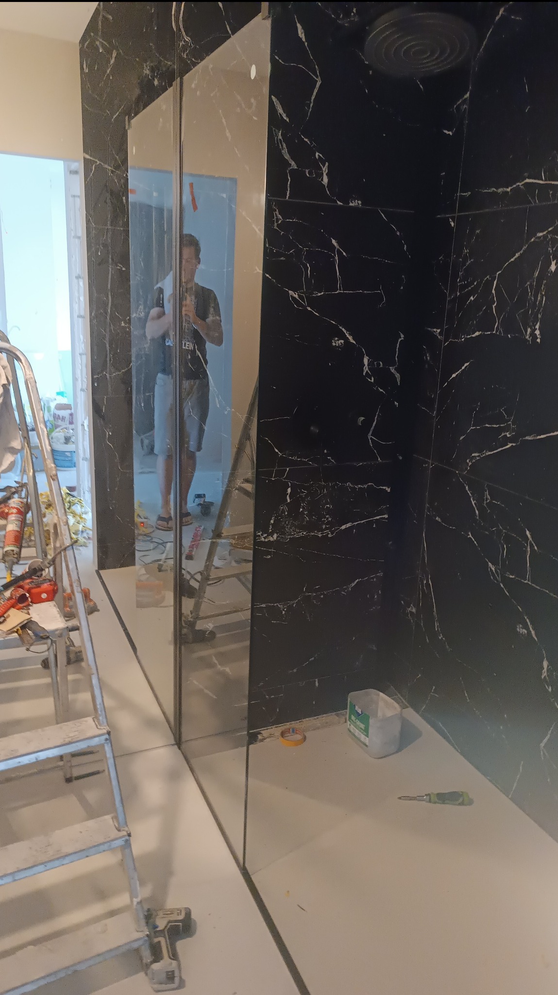 Częściowo wykończona łazienka z czarnymi marmurkowymi płytkami i szklaną kabiną prysznicową, widoczna drabina i narzędzia sugerują trwający proces.