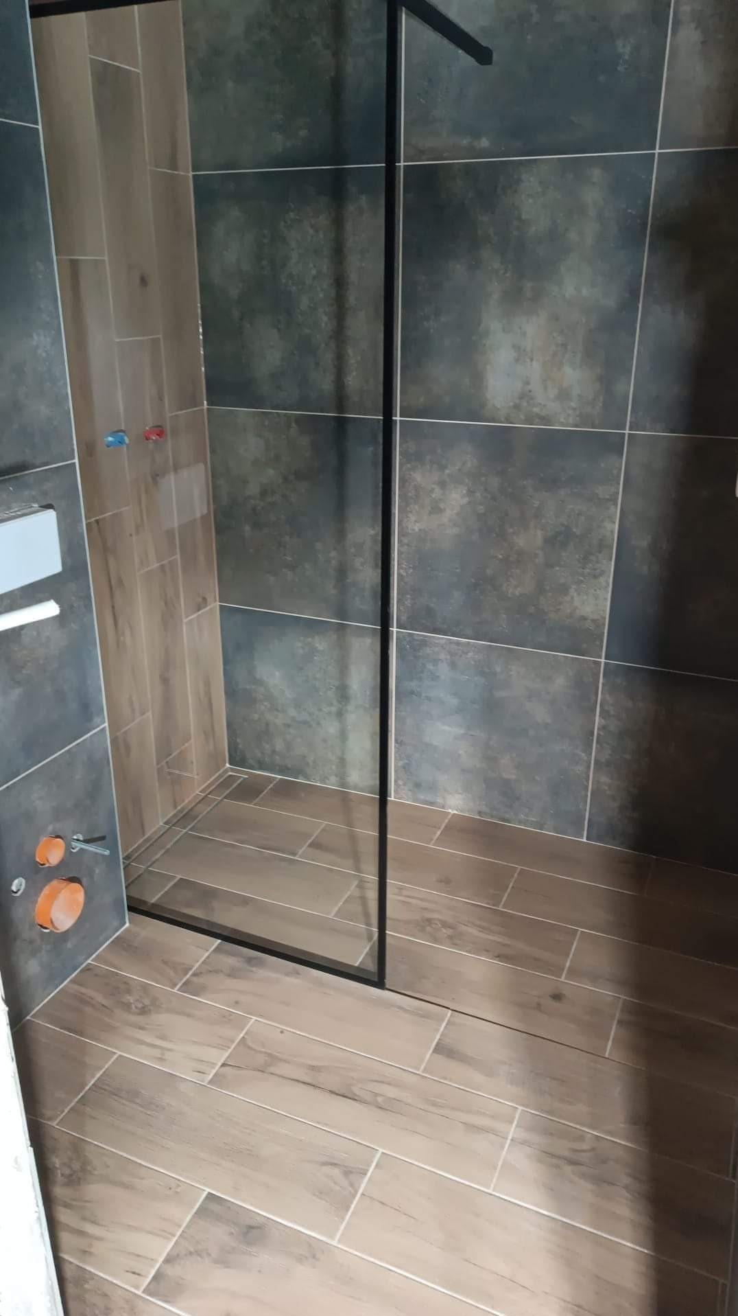 Nowoczesna kabina prysznicowa typu walk-in z czarną ramą, wykończona płytkami w odcieniach szarości i drewna, widoczne podłączenia wodne i odpływ liniowy.