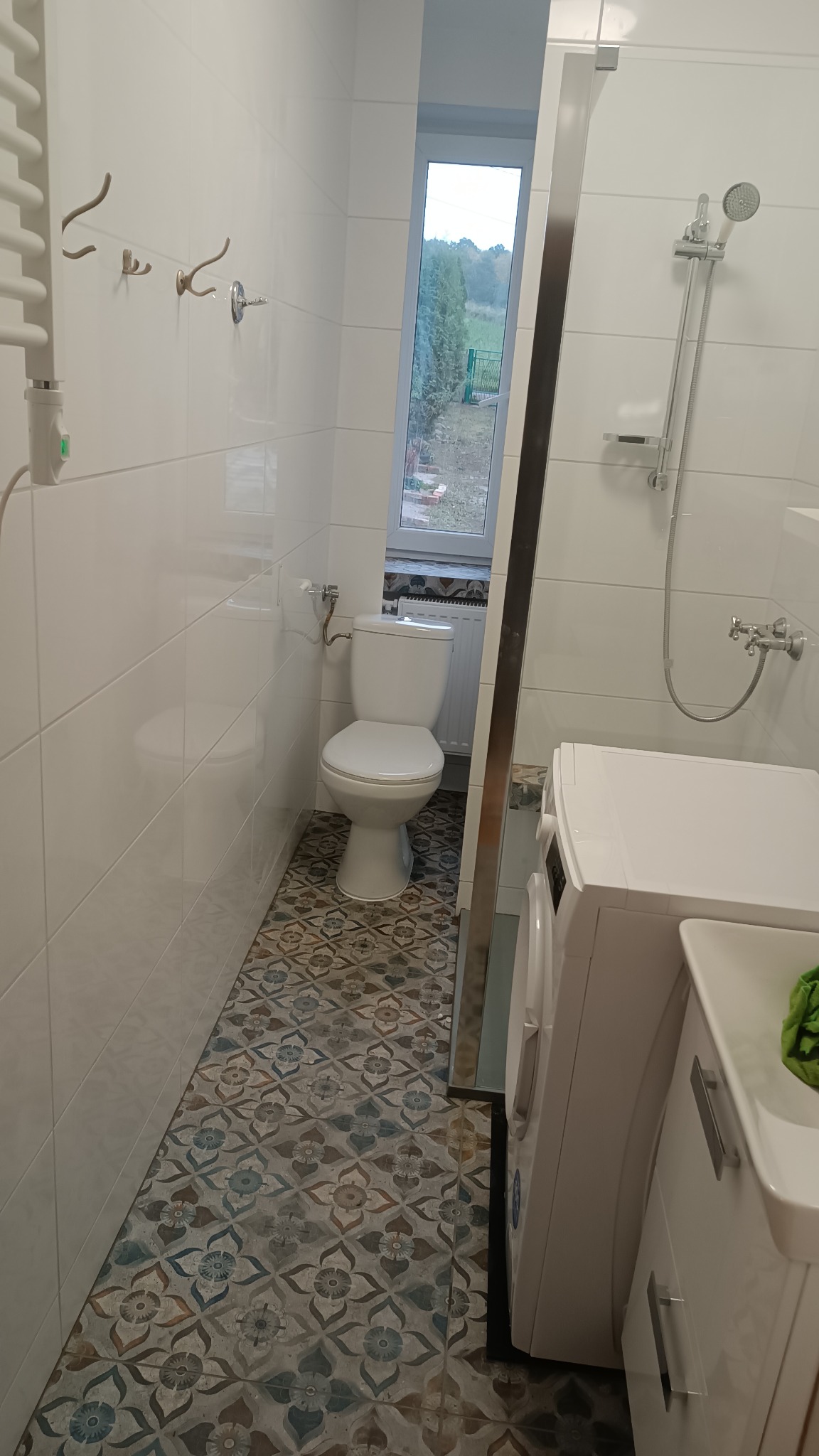 Wąska łazienka z białą armaturą, wzorzystą podłogą i oknem z widokiem na zieleń.