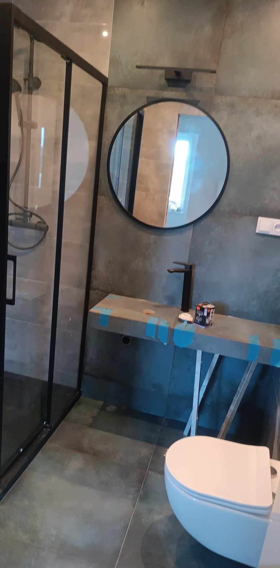 Nowoczesna łazienka w trakcie wykańczania: szara glazura, okrągłe lustro w czarnej ramie, czarna bateria umywalkowa, kabina prysznicowa z czarnymi profilami, umywalka zabezpieczona niebieską taśmą...