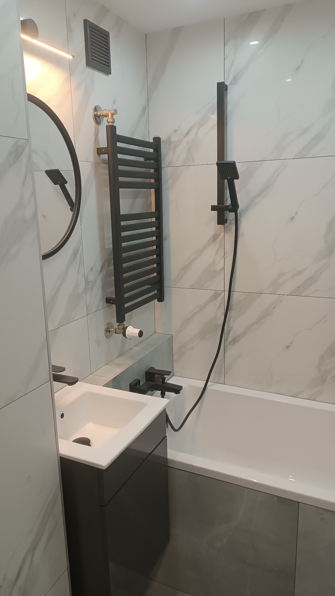 Nowoczesna łazienka z marmurowymi płytkami, czarną armaturą, grzejnikiem drabinkowym i okrągłym lustrem z podświetleniem LED.