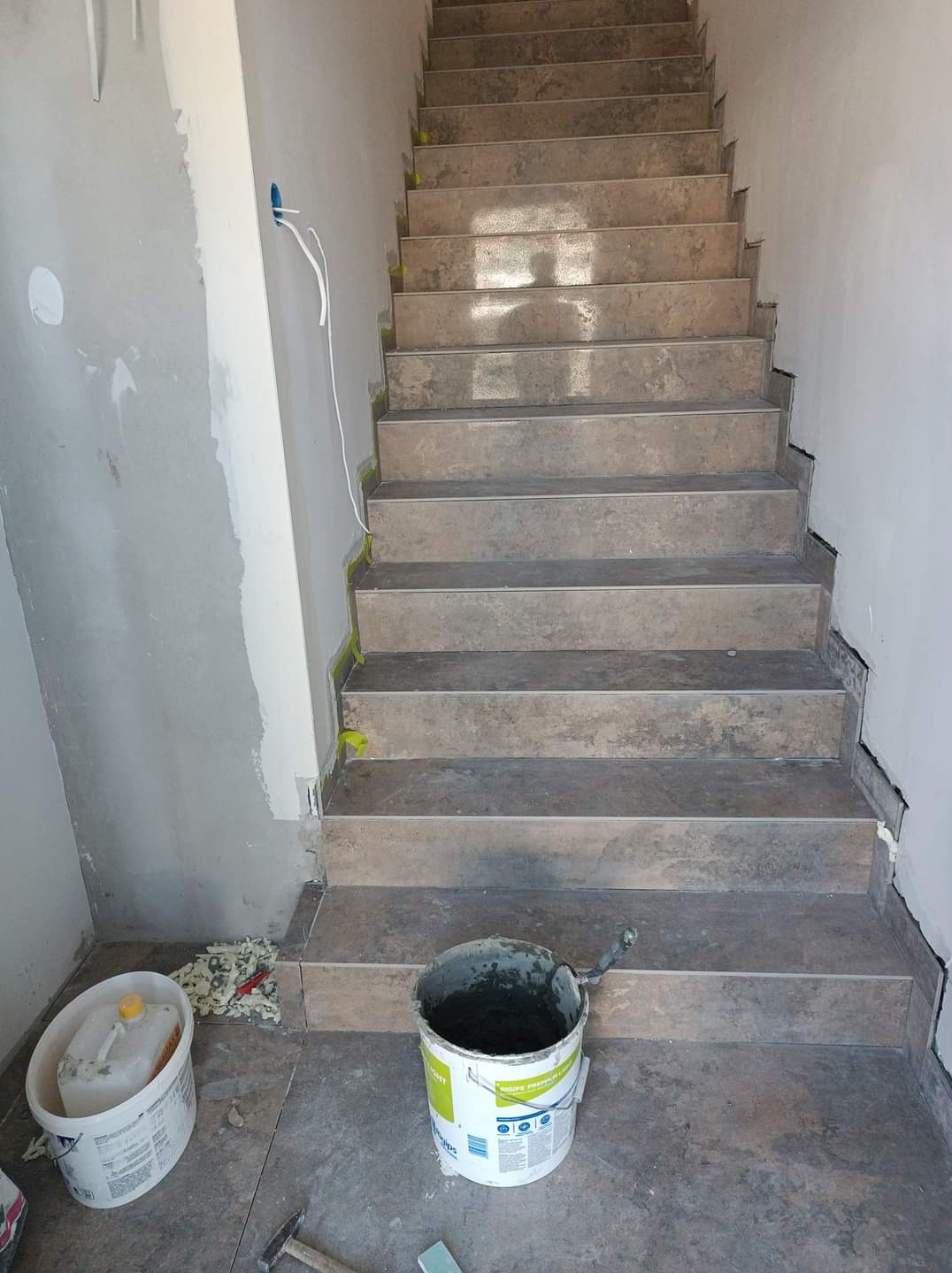 Montaż płytek na schodach w trakcie remontu, widoczne wiadro z zaprawą, poziomica i młotek na podłodze, zabezpieczenia taśmą malarską.