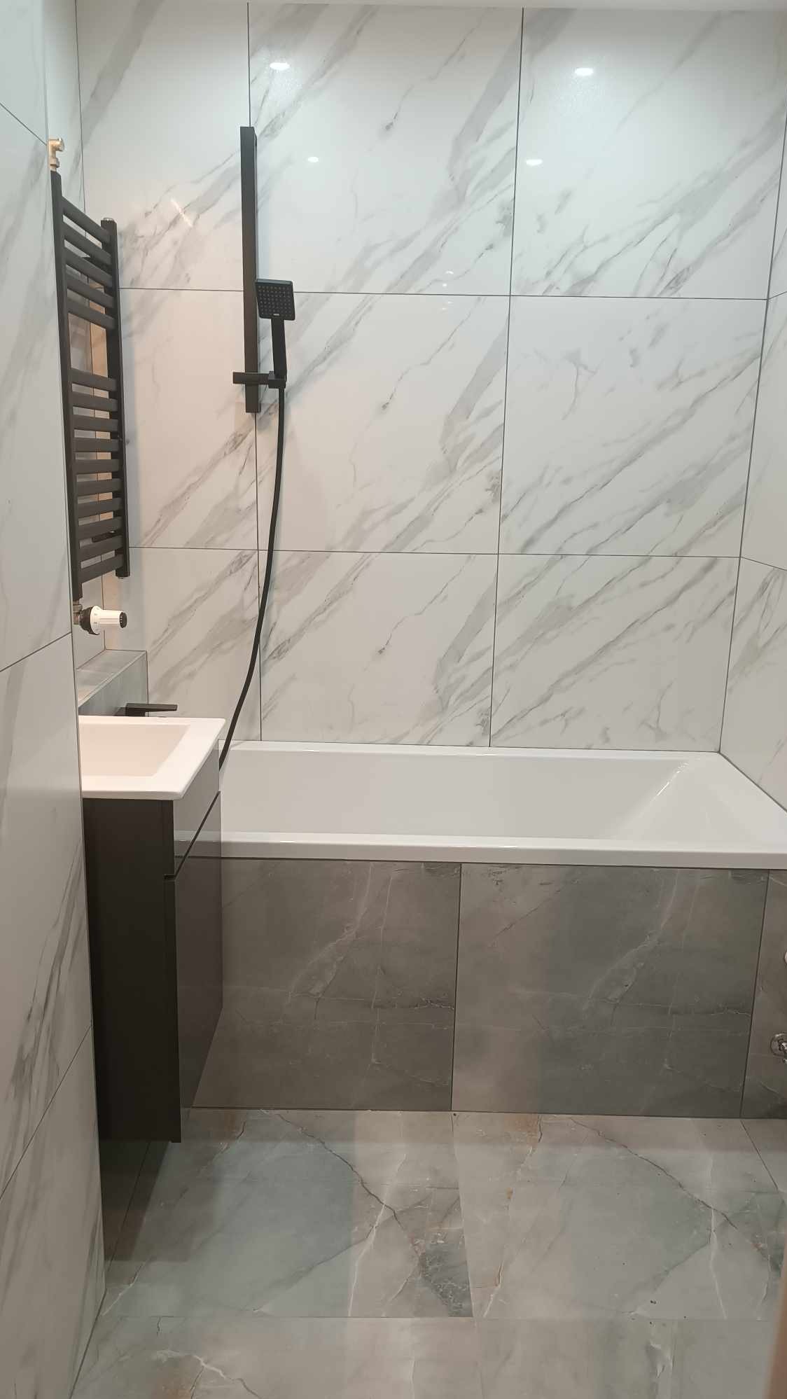 Nowoczesna łazienka z wanną obudowaną szarymi płytkami, kabiną prysznicową w kolorze czarnym, umywalką z szarą szafką i grzejnikiem drabinkowym.