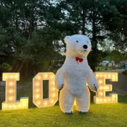 Duży, biały, pluszowy miś z czerwoną muszką stoi na trawie przed podświetlanym napisem LOVE.