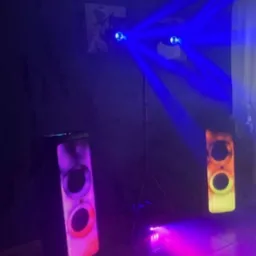 Dwa głośniki JBL PartyBox z podświetleniem LED, oświetlenie sceniczne z niebieskimi promieniami, ciemne tło, płytki podłogowe.