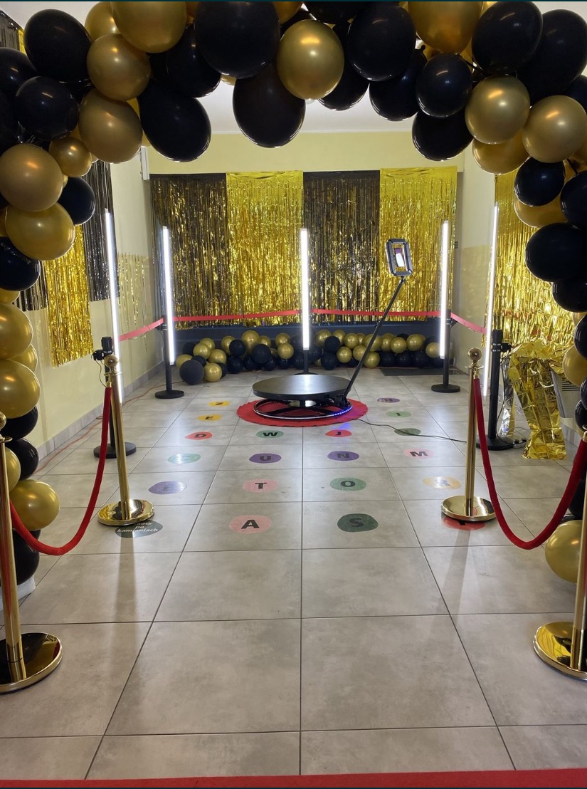 Rozstawiona obrotowa platforma do nagrywania filmów 360 stopni, otoczona złotymi i czarnymi balonami oraz złotymi frędzlami na ścianie, wyznaczona czerwonymi słupkami z linami, na podłodze...
