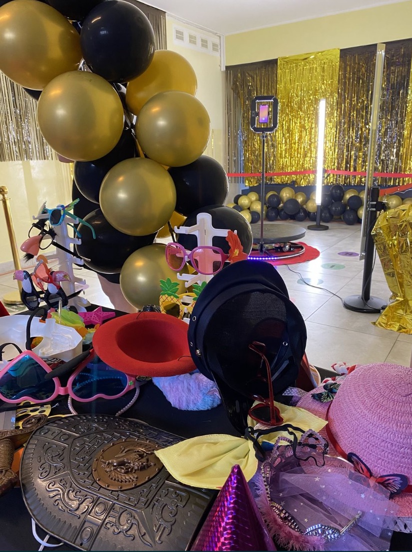 Przygotowane rekwizyty, takie jak kapelusze, okulary i peruki, na stole przed fotobudką z tłem w kolorze złotym i czarnymi balonami, w Redzie. Widoczny automat do zdjęć na obrotowej platformie.