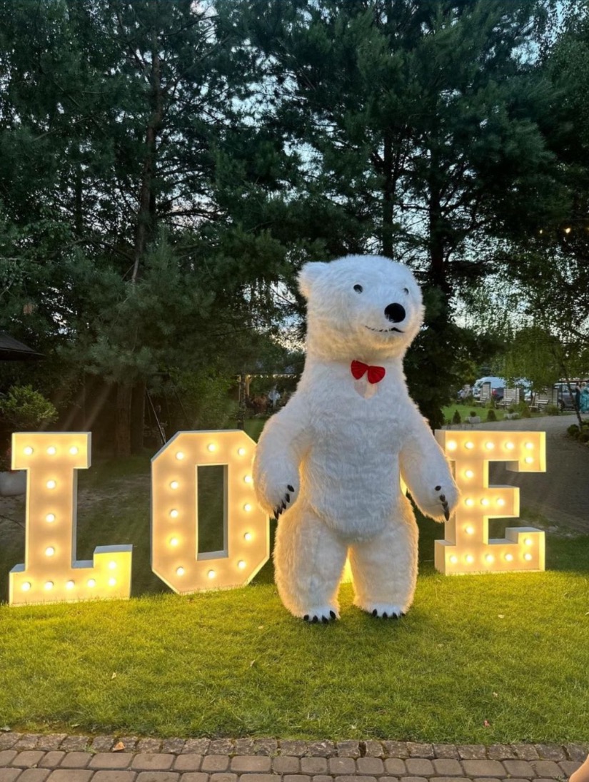 Duży, biały, pluszowy miś z czerwoną muszką stoi na trawie przed podświetlanym napisem LOVE.