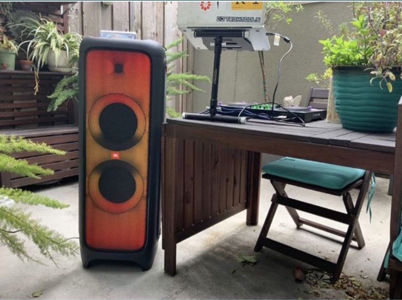 Głośnik JBL Partybox obok stołu DJ-skiego z laptopem i gramofonami na zewnątrz, otoczenie rośliny doniczkowe i drewniane ogrodzenie.