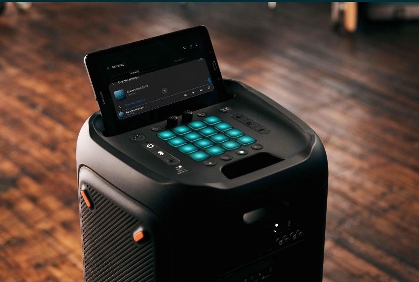 Czarny głośnik imprezowy z podświetlanymi na niebiesko przyciskami i tabletem Samsung wyświetlającym listę utworów, ustawiony na drewnianej podłodze.