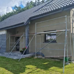 Dom jednorodzinny w trakcie prac wykończeniowych elewacji, widoczne rusztowania, ocieplona ściana w kolorze szarym, fragment wykończony kamieniem elewacyjnym, dachówka ceramiczna w kolorze...
