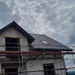 Lox-group - Dom w trakcie budowy z częściowo ukończonym dachem z dachówką, widoczne rusztowania i pracownik przy ogrodzeniu, pochmurne niebo w tle.